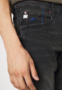 Primer plano de una mano descansando cerca del bolsillo delantero de unos jeans negros desgastados con costuras azules y una pequeña etiqueta de tela blanca.