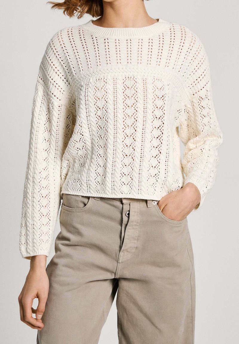 Strickpullover in Off-White mit Lochmustern, Rundhalsausschnitt, überschnittenen Schultern und einer verkürzten Silhouette. Kombiniert mit beigen Hosen.