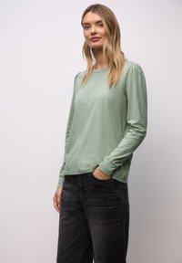 Femme aux cheveux blonds portant une chemise à manches longues vert clair et un jean gris foncé, debout avec une main dans la poche devant un fond uni.