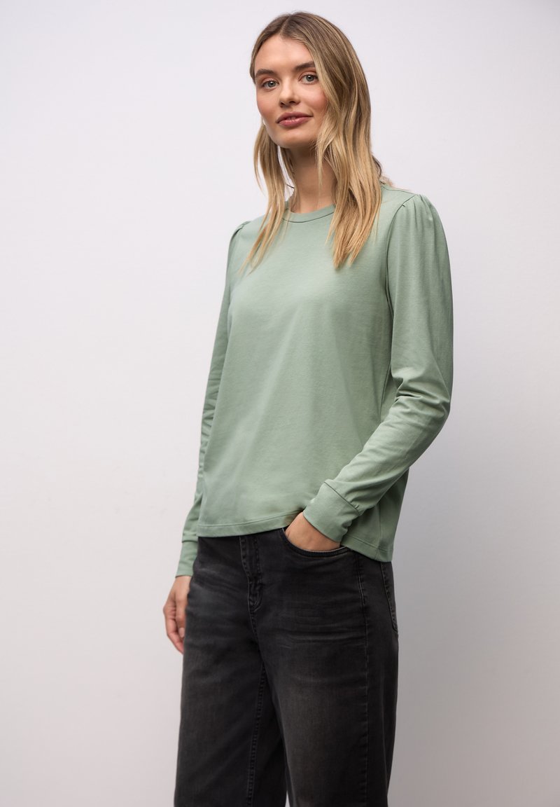 Femme aux cheveux blonds portant une chemise à manches longues vert clair et un jean gris foncé, debout avec une main dans la poche devant un fond uni.