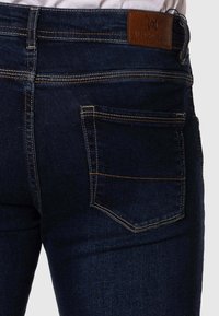 Vaqueros de mezclilla azul oscuro con un bolsillo trasero, costuras amarillas contrastantes y una etiqueta de cuero marrón con el nombre de la marca en la cintura.