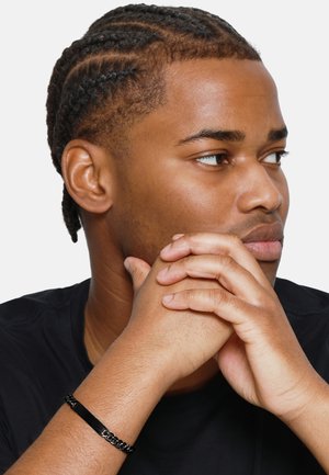 Braccialetto a catena nera con un accento rettangolare piatto, indossato su un polso. La persona ha i capelli intrecciati e tiene il mento appoggiato sulle mani.