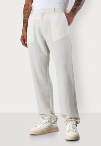 A person wears light-colored, linen blend pants and white sneakers, with hands in pockets, against a neutral background.