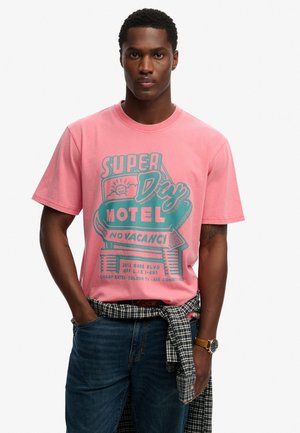 Joven con una camiseta rosa con un gráfico azul verdoso que dice "Super Day Motel No Vacancy", jeans azules y una camisa a cuadros atada alrededor de su cintura.