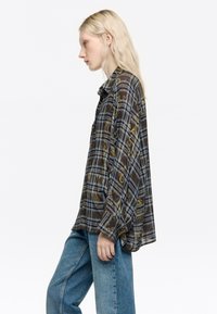 Camisa de cuadros marrón y azul de manga larga con patrones de hojas, corte relajado y cuello tipo solapa. Combinada con jeans azules de tiro alto.