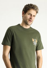 Camiseta de algodón verde oliva con mangas cortas, cuello redondo y un gráfico frontal que presenta un vehículo vintage y árboles en color beige.