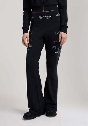 Person iført sorte vidde bukser med hvide kløer og "Ed Hardy" tekstdesign, kombineret med sorte sko og en kort sort jakke.