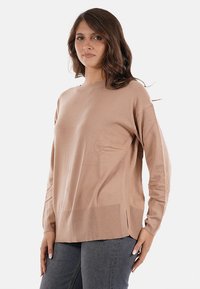 Donna con capelli castani mossi, che indossa un maglione beige a maniche lunghe e larghe, insieme a jeans grigio scuro, in piedi su uno sfondo neutro.