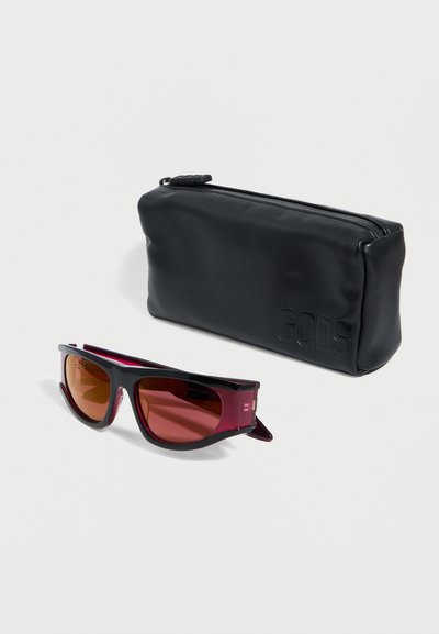 Μαύρα και σκουροκόκκινα ορθογώνια γυαλιά ηλίου με φακούς διαβαθμισμένου τόνου δίπλα σε μαύρη θήκη με φερμουάρ, με ανάγλυφο το λογότυπο "GCDS".