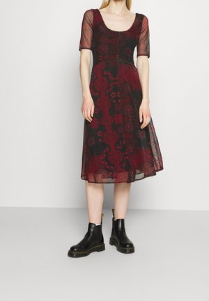 Kobieta ubrana w czarną i ciemnoczerwoną sukienkę o długości do kolan z przeźroczystymi rękawami do łokcia oraz czarne, masywne botki do kostki.