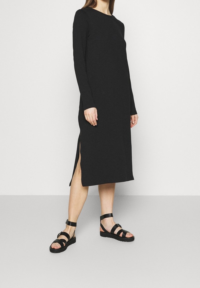Femme portant une robe midi noire à manches longues avec une fente latérale et des sandales plates noires à brides, debout sur un sol gris.