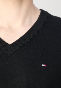 Gros plan d'une personne portant un pull noir à col en V avec un petit logo rectangulaire rouge, blanc et bleu sur la poitrine.