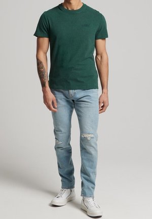 Grünes Baumwoll-T-Shirt mit Rundhalsausschnitt, kombiniert mit hellblauen, verwaschenen Jeans und weißen Turnschuhen. Das Model steht vor einem neutralen Hintergrund.