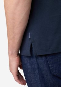 Marineblauwe katoenen t-shirt met zijsplitten, voorzien van een klein merklabel bij de zoom. Draagt samen met donkere denim jeans, met de hand ontspannen aan de zij.