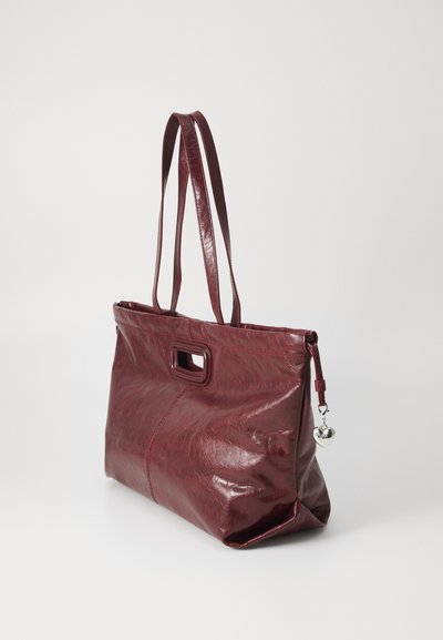 Red leather tote bag with a smooth texture, featuring two shoulder straps and a heart-shaped charm. The bag has a rectangular shape and a cut-out handle.
