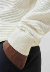 Pull en tricot blanc à côtes avec un tissu texturé et un poignet froncé. Tissu sombre visible sous le pull.
