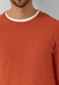 Man draagt een gebrande oranje trui met ronde hals over een wit ondershirt, waarbij de hals, een deel van de baard en de bovenste romp zichtbaar zijn tegen een grijze achtergrond.