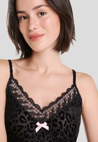 Camiseta sin mangas negra con estampado de leopardo, con delicados bordes de encaje negro y un lazo rosa claro en el centro. Tirantes delgados ajustables. Textura suave.