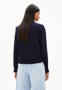 Marineblaue Strickweste mit strukturierter Oberfläche, ärmelloses Design, kombiniert mit einem dunklen Langarmhemd und hellblauen High-Waist-Jeans.