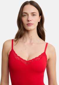 Frau mit langem braunem Haar trägt ein rotes Camisole mit Spitzenbesatz und blickt nach vorne vor einem einfachen weißen Hintergrund.