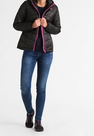 Personne portant une doudoune noire avec une fermeture éclair rose, un jean bleu foncé et des baskets noires avec des accents violets, debout devant un fond blanc.