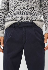 Donkerblauwe katoenen broek met een knoopsluiting, voorplooien en een gladde textuur. Een gebreid trui met patroon is zichtbaar bij de taille.
