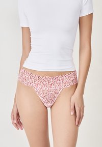 Femme portant un haut blanc à manches courtes et une culotte en dentelle rose avec un motif de cœurs rouges, se tenant devant un fond clair.