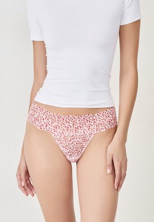 Femme portant un haut blanc à manches courtes et une culotte en dentelle rose avec un motif de cœurs rouges, se tenant devant un fond clair.