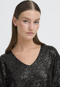 Mustassa helmikirjailulla varustettu paita, jossa on V-muotoinen kaula-aukko ja lyhyet hihat. Paidassa on teksturoidut pinnat ja kimaltavat yksityiskohdat.