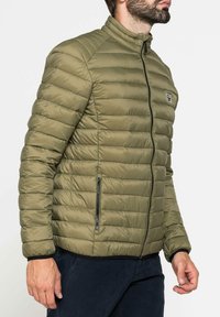 Giacca a vento verde oliva con texture trapuntata, colletto alto, zip frontale e tasche laterali. Presenta bordi neri lungo l'orlo e la zip.