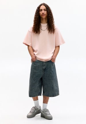 Tricou roz deschis, supradimensionat, pantaloni scurți din denim până la genunchi, adidași gri cu mai multe curele, coliere din argint în straturi, păr creț.