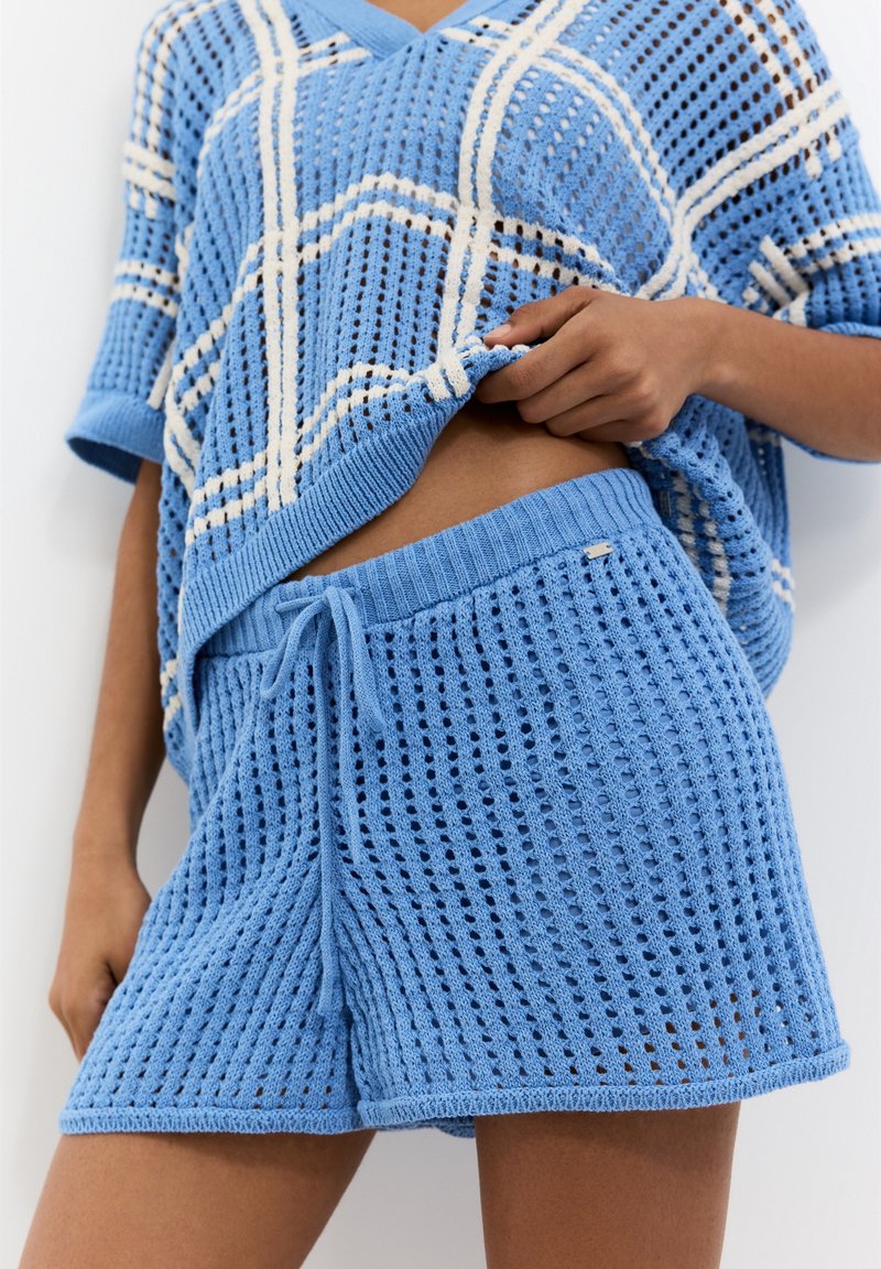 Conjunto de loungewear de color azul claro, con un top de tejido suelto y patrón abierto, y pantalones cortos a juego con cintura elástica y cordón ajustable.