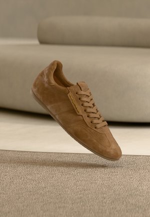 Brązowy zamszowy trampek z zaokrąglonym noskiem, sznurowanym przodem, złotym logo oraz teksturowaną podeszwą, zawieszony nad neutralnym tłem.