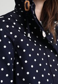 Marinblå jacka med vita prickar, försedd med huva, justerbar dragsko och kontrasterande knapppdetaljer. Sl ät tygstruktur.