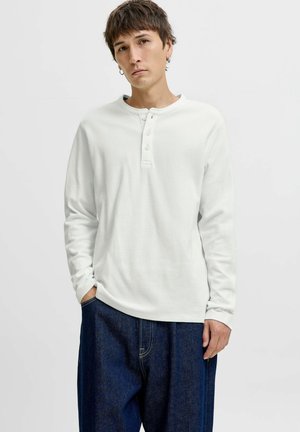 Young man wearing a white long-sleeve henley shirt with three buttons and loose dark blue denim jeans, standing with one hand in pocket.