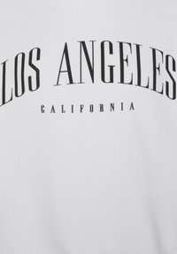 Felpa bianca con testo in grassetto nero che recita "LOS ANGELES" arcuato sopra "CALIFORNIA", caratterizzata da una texture liscia e morbida.