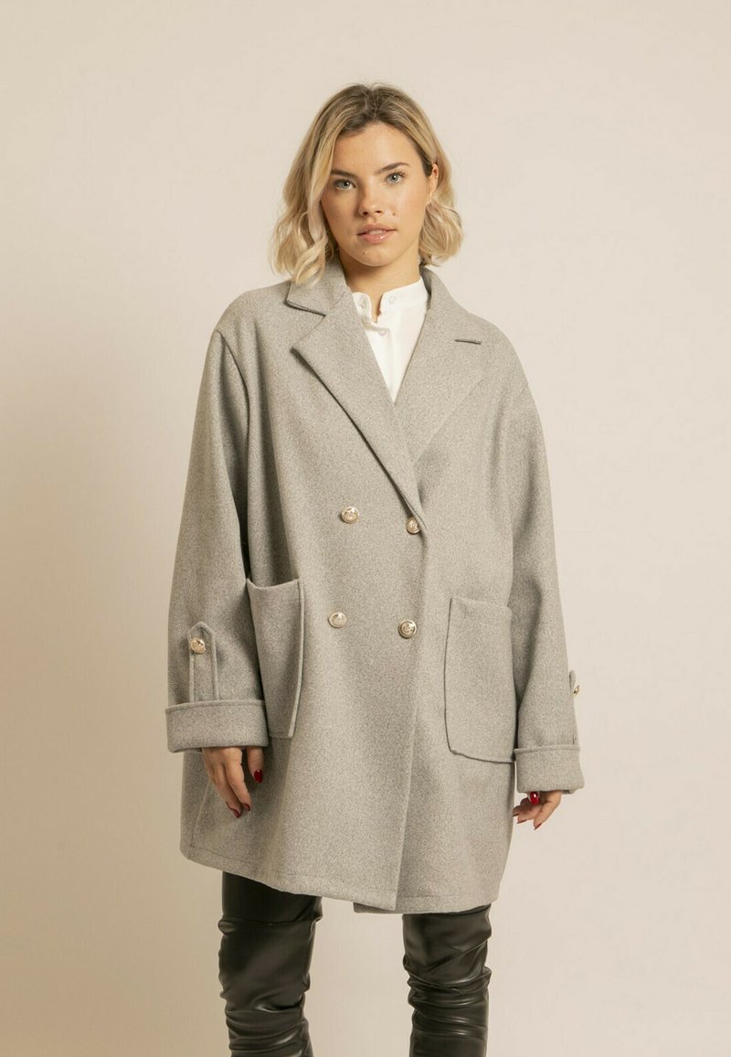 Manteau gris à double boutonnage avec de grandes poches, en tissu texturé. Avec des boutons dorés et des poignets larges, associé à un pantalon en cuir.