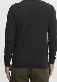 Pull noir à manches longues, poignets et ourlet côtelés, texture lisse, avec un empiècement arrondi dans le dos, associé à un pantalon vert.