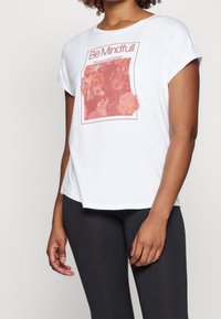 Biała koszulka z krótkim rękawem z różowym kwiatowym wzorem i tekstem. Luźny krój z okrągłym dekoltem i prostym dołem. Noszona z czarnymi legginsami.