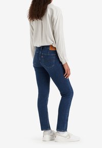 Vaqueros azul oscuro de corte ajustado, con costuras reforzadas y un parche de cuero en la parte trasera de la cintura; combinados con una camisa de manga larga de rayas claras.