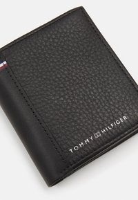 Černá kožená peněženka s texturovaným povrchem, s bílým logem a barevným pruhovaným akcentem na jedné straně. Kompaktní a obdélníkový design.