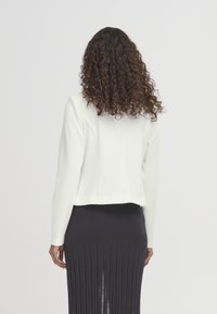 Veste blanche courte à manches longues, texture lisse, épaules arrondies et coutures latérales. Associée à une jupe noire à franges.