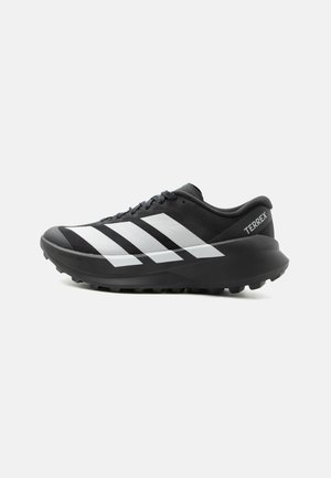 Čierno-biela trailová bežecká obuv Adidas Terrex s hrubou podrážkou a drážkovanou vonkajšou podrážkou, bočný pohľad na jednofarebnom svetlom pozadí.