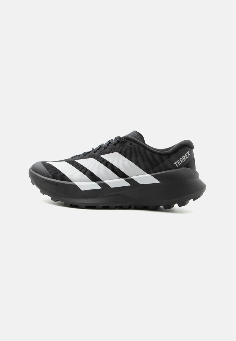 Chaussure de trail running Adidas Terrex noire et blanche avec une semelle épaisse et une semelle extérieure à crampons, vue de côté sur un fond clair uni.