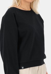 Musta puuvillapaita, jossa on pyöreä kaula-aukko, ribbireunat hihansuissa ja helmassa. Alareunassa pieni merkkilogo. Pehmeä rakenne.