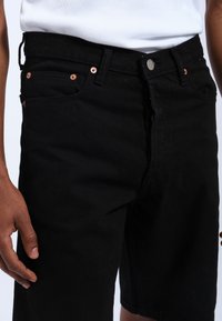 Shorts de mezclilla negra con corte recto, que cuentan con remaches de cobre, bolsillos delanteros y cierre de botón. Textura suave con un diseño clásico.
