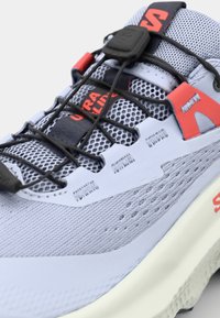 Chaussure de course gris clair avec une tige en maille texturée, accents noirs et rouges, système de laçage flexible et semelle intermédiaire blanche rembourrée.