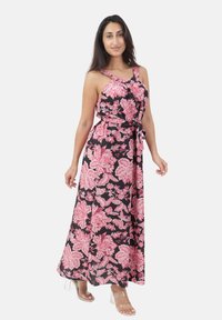 Donna che indossa un vestito maxi nero senza maniche con stampa floreale rosa e sandali con tacco trasparenti, in piedi davanti a uno sfondo bianco.