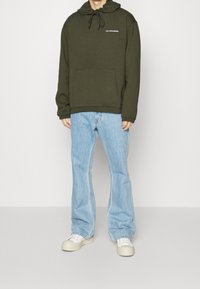 Felpa verde oliva con tasca a marsupio frontale, cappuccio con cordino e testo bianco. Abbinata a jeans svasati blu chiaro e sneakers color crema.