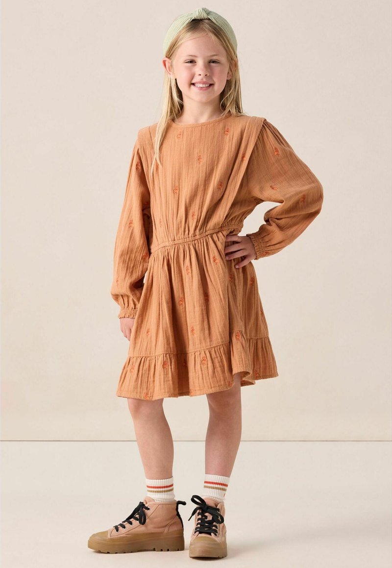 Robe en coton marron à manches longues, taille froncée, ourlet volanté et détails brodés orange. Portée avec des baskets beige et des chaussettes rayées.